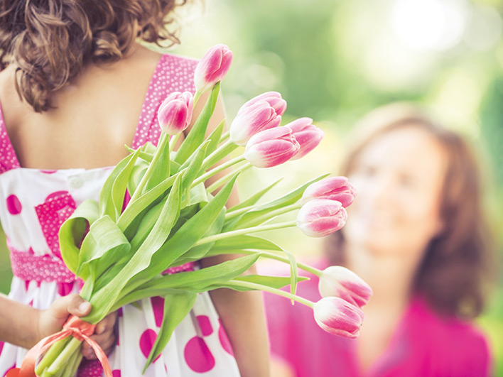 Young girl with tulips behind her back with mother blurred in the distance